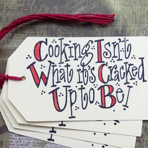 May include: Cream-coloured gift tags with the text "Cooking Isn't What It's Cracked Up To Be!" in black and red. A red twine is attached to the top tag. The tags are stacked.