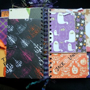 May include: A Halloween-themed journal with a spiral binding, featuring pages with ghost, skeleton, and pumpkin patterns. Includes orange and purple ribbons, and the text "Jack-O-Land".