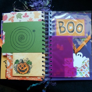 May include: A Halloween-themed journal with a spiral binding, featuring decorative pages. The journal includes a green card with a spiderweb design, a pumpkin illustration, and a page with the word "BOO". The pages are decorated with autumn leaves and a ghost cutout.