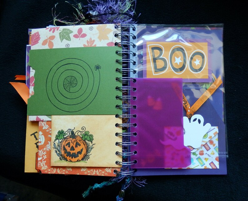 May include: A Halloween-themed journal with a spiral binding, featuring decorative pages. The journal includes a green card with a spiderweb design, a pumpkin illustration, and a page with the word "BOO". The pages are decorated with autumn leaves and a ghost cutout.