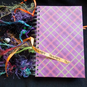 May include: A spiral-bound journal with a purple plaid cover. The journal is decorated with colourful ribbons and decorative strands in shades of orange, purple, green, and red. A ribbon with the words "Trick-or-Treat" is attached.