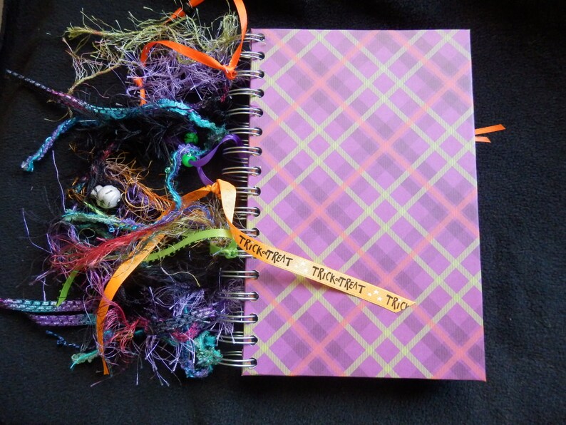 May include: A spiral-bound journal with a purple plaid cover. The journal is decorated with colourful ribbons and decorative strands in shades of orange, purple, green, and red. A ribbon with the words "Trick-or-Treat" is attached.