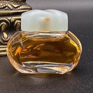 May include: A small, clear glass perfume bottle with a light blue, frosted stopper. The bottle is filled with a golden-coloured liquid. The bottle has an oval shape with a faceted design. The background includes a decorative gold object and faux pearls.