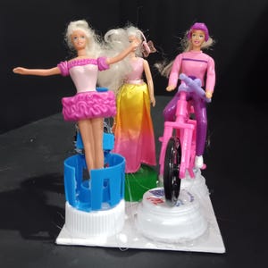 May include: Three miniature dolls, each with blonde hair, are displayed on a white base. One doll wears a pink ballet outfit, another a yellow and pink dress, and the third doll is riding a pink bicycle. The dolls are placed on bottle caps.
