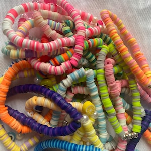 Könnte beinhalten: Eine Sammlung bunter Armbänder aus flachen, runden Polymer-Ton-Perlen in verschiedenen Farben wie Neonpink, Orange, Gelb, Blau, Lila und Weiß, akzentuiert mit kleinen weißen Perlen und einem gelben Zitronenscheiben-Anhänger.