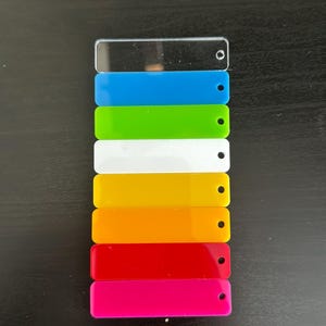 May include: A selection of brightly coloured acrylic keyrings in a rainbow sequence: clear, blue, green, white, yellow, orange, red, and pink. Each keyring is rectangular with a hole for attachment. A yellow keyring with "LOV3 YOU" is also visible.