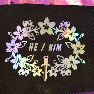 May include: Black fabric patch with a white iridescent design of a floral wreath with a dagger in the center. The text "HE/HIM" is inside the wreath.
