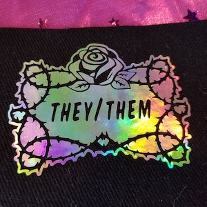 Pode incluir: Um patch holográfico para passar a ferro com um contorno preto de uma rosa com espinhos e o texto "THEY/THEM" no interior.