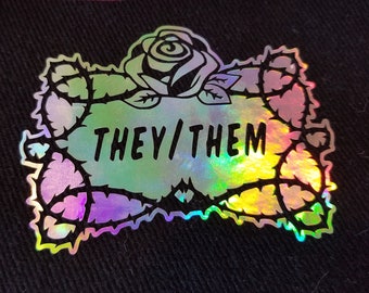 May include: A holographic iron-on patch with a black outline of a rose with thorns and the text "THEY/THEM" inside.