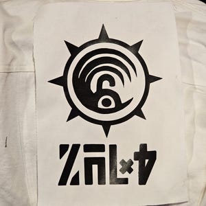 以下が含まれることがあります： 背中に大きな黒いグラフィックパッチが付いた白いデニムジャケット。パッチは、様式化された波模様と太字のブロック文字で「ZAL+4」というテキストを備えた円形デザインが特徴です。