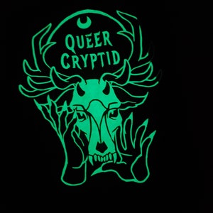 May include: A green glow-in-the-dark sticker with a black and white illustration of a creature with antlers, hands covering its face, and the text "QUEER CRYPTID".