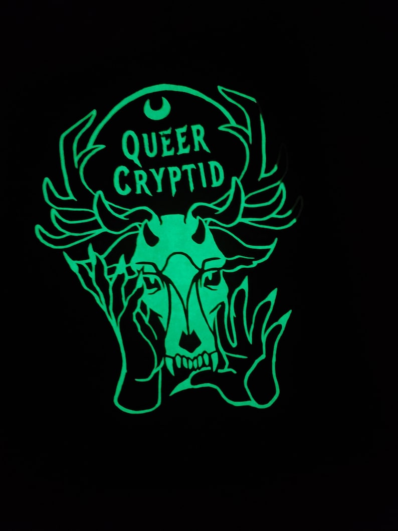 May include: A green glow-in-the-dark sticker with a black and white illustration of a creature with antlers, hands covering its face, and the text "QUEER CRYPTID".