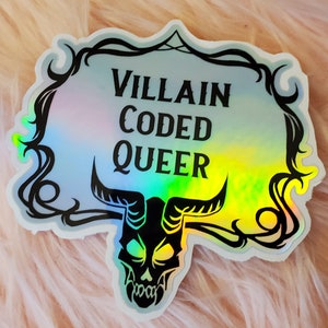 May include: A holographic sticker with the text "VILLAIN CODED QUEER" in black, set against a light blue and rainbow background. The sticker features a black skull with horns and decorative swirls.