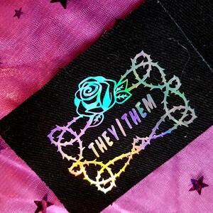 May include: A black fabric patch with a holographic rose and thorns design. The text "THEY/THEM" is inside the thorns.