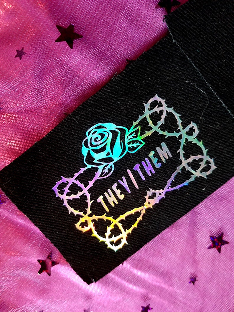 May include: A black fabric patch with a holographic rose and thorns design. The text "THEY/THEM" is inside the thorns.
