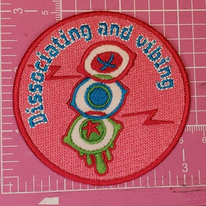 May include: Pink embroidered patch with the text "Dissociating and vibing" in white. The patch features three stylized eyes with green, blue, and red accents.