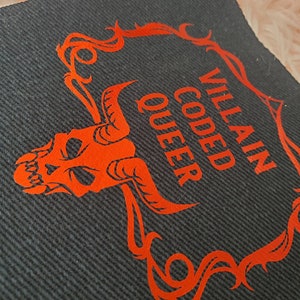 Peut inclure: Un patch en tissu noir avec un cr&acirc;ne rouge et un cadre orn&eacute;. Le texte "Villain Coded Queer" est &eacute;crit en rouge.