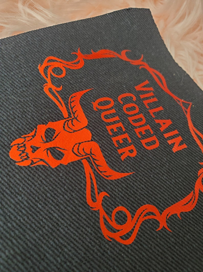 Peut inclure: Un patch en tissu noir avec un cr&acirc;ne rouge et un cadre orn&eacute;. Le texte "Villain Coded Queer" est &eacute;crit en rouge.