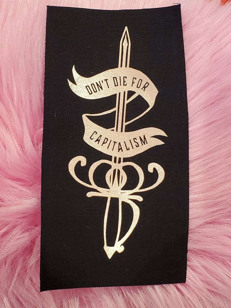 May include: Black fabric patch with gold embroidery of a sword with a banner that reads "Don't Die For Capitalism".