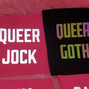 Może przedstawiać: Dwa łatki z tkaniny z tekstem "QUEER JOCK" w kolorze białym na czerwonym tle i "QUEER GOTH" w tęczowych, opalizujących literach na czarnym tle.