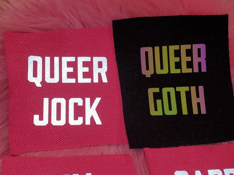 Może przedstawiać: Dwa łatki z tkaniny z tekstem "QUEER JOCK" w kolorze białym na czerwonym tle i "QUEER GOTH" w tęczowych, opalizujących literach na czarnym tle.