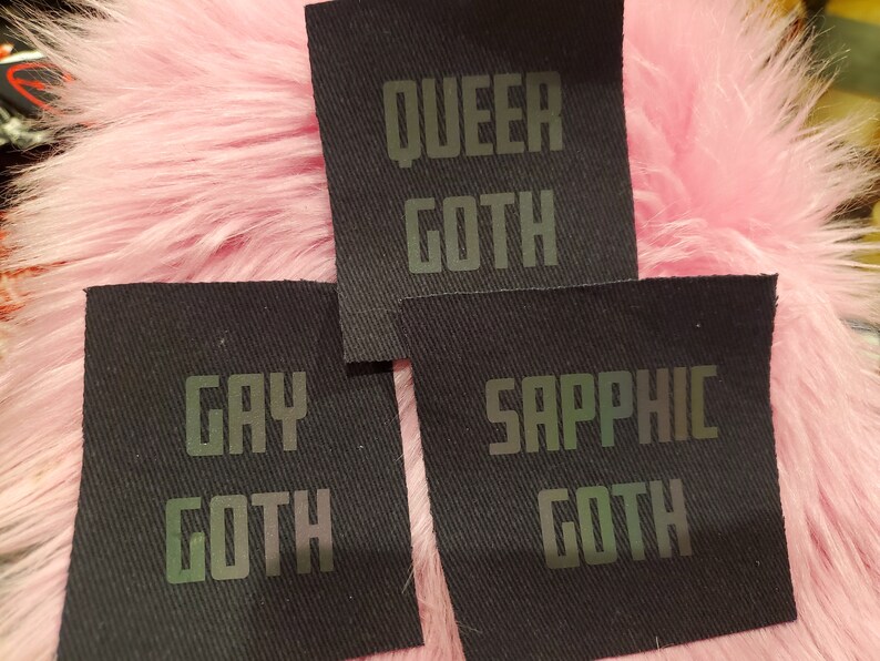 Może przedstawiać: Trzy czarne łatki z tkaniny z iryzującym zielonym tekstem. Łatki głosiły "Queer Goth", "Gay Goth" i "Sapphic Goth".