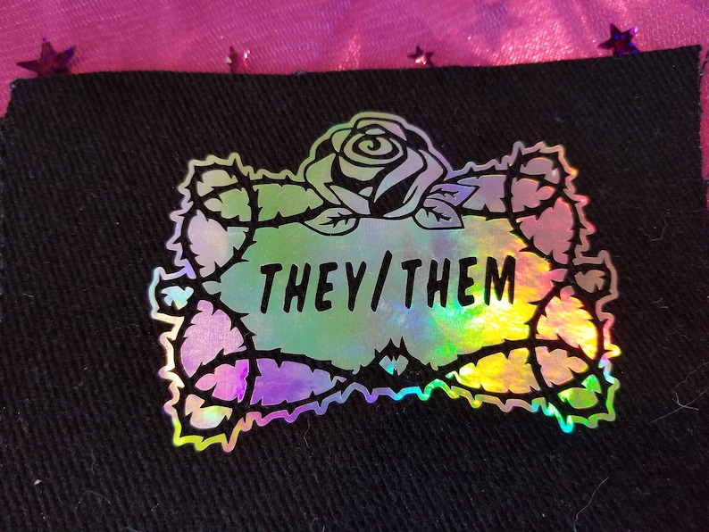 May include: A holographic iron-on patch featuring a black outline of a rose with thorns and the text "THEY/THEM" inside.