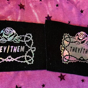 May include: Two black fabric patches with a holographic rose and thorns design. The text "THEY/THEM" is inside the design.