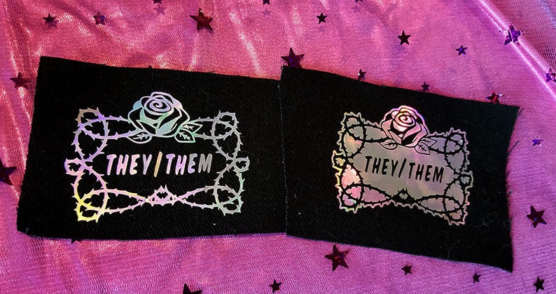 May include: Two black fabric patches with a holographic rose and thorns design. The text "THEY/THEM" is inside the design.