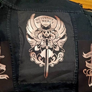 May include: A black denim jacket with three patches. The center patch features a winged figure holding a sword with the text "No Justice No Mercy". The left patch features a sword with the text "Fortune Favors the Bold". The right patch features a sword with the text "Don't Die For Capitalism".