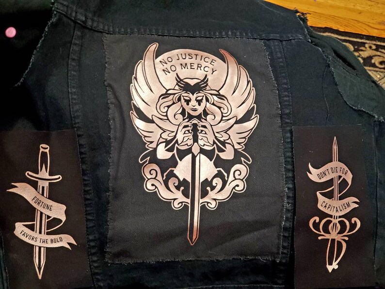 May include: A black denim jacket with three patches. The center patch features a winged figure holding a sword with the text "No Justice No Mercy". The left patch features a sword with the text "Fortune Favors the Bold". The right patch features a sword with the text "Don't Die For Capitalism".