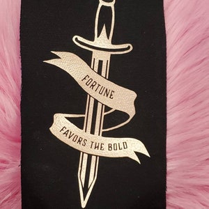 May include: A black fabric patch with a gold foil design of a dagger with a banner that reads "Fortune Favors the Bold".