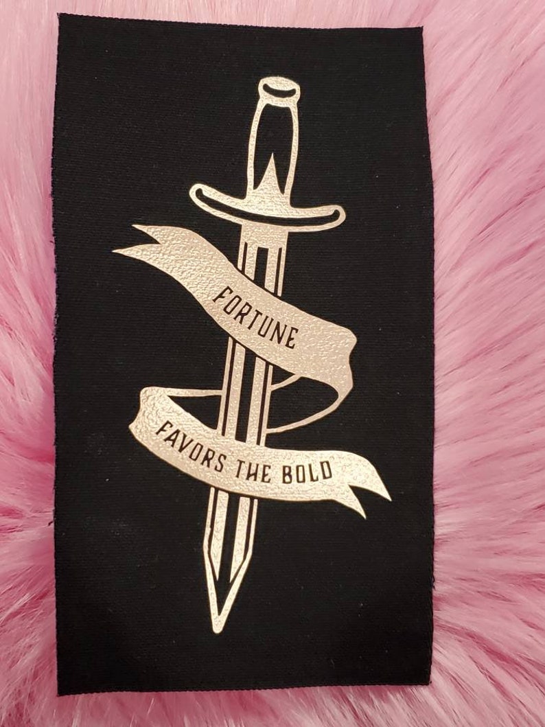 May include: A black fabric patch with a gold foil design of a dagger with a banner that reads "Fortune Favors the Bold".