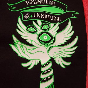 May include: Black patch with a green and white design. The design features a pair of wings, three eyes, a hand, and a stack of three discs. The text "Supernatural Not Unnatural" is written in a banner above the design.