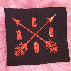 Op de afbeelding: Een zwarte stoffen patch met een rood geborduurd ontwerp. Twee gekruiste pijlen met een gestileerde "C.A.B" in het midden.
