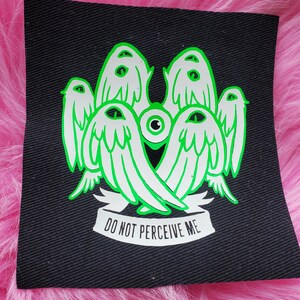 May include: Black patch with a green and white design of five winged figures with eyes. The figures are arranged in a circle around a central eye. The text "DO NOT PERCEIVE ME" is printed below the figures.
