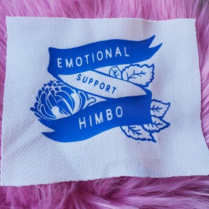 May include: A white fabric patch with blue text and a blue floral design. The text reads "Emotional Support Himbo".