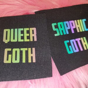 Może przedstawiać: Dwa czarne łatki z tkaniny z tęczowymi, opalizującymi napisami. Pierwsza łatka głosi "Queer Goth", a druga "Sapphic Goth".