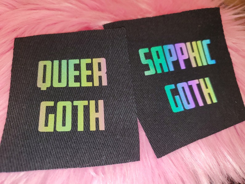 Może przedstawiać: Dwa czarne łatki z tkaniny z tęczowymi, opalizującymi napisami. Pierwsza łatka głosi "Queer Goth", a druga "Sapphic Goth".