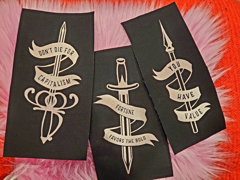 May include: Three black fabric patches with gold embroidery. The patches feature swords and phrases: "Don't Die For Capitalism", "Fortune Favors The Bold", and "You Have Value".
