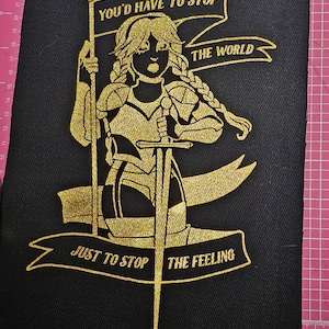 May include: A gold glitter iron-on patch with a female warrior holding a sword and a banner that reads "You'd have to stop the world just to stop the feeling."