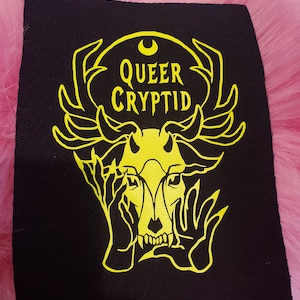 May include: A black fabric patch with a yellow design of a skull with antlers and hands covering its face. The text "QUEER CRYPTID" is above the skull.