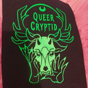 May include: Green and black embroidered patch with a stylized creature with antlers and hands over its face. The text "QUEER CRYPTID" is above the creature.