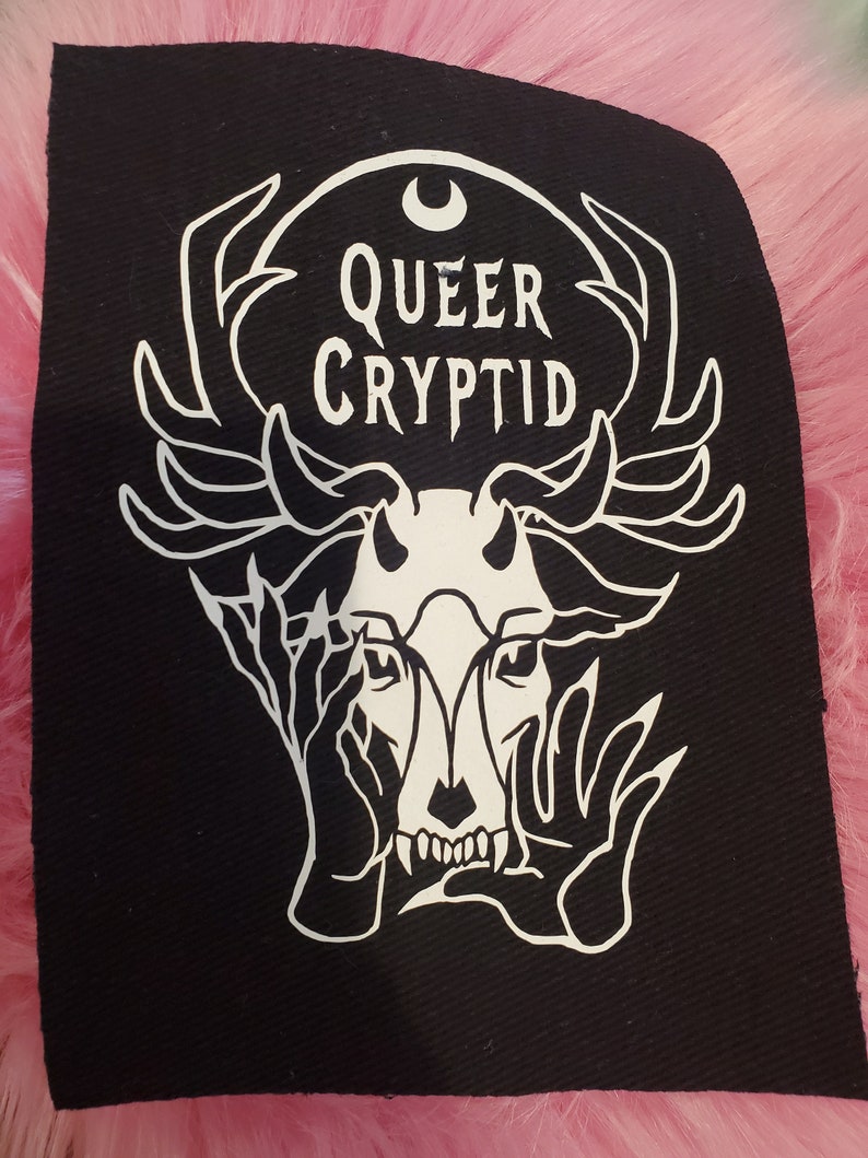 May include: Black and white iron-on patch with a stylized image of a deer skull with antlers, hands covering its eyes, and the text "QUEER CRYPTID" above it.