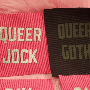 May include: Two pink fabric patches with silver lettering that reads "QUEER JOCK" and "QUEER GOTH".