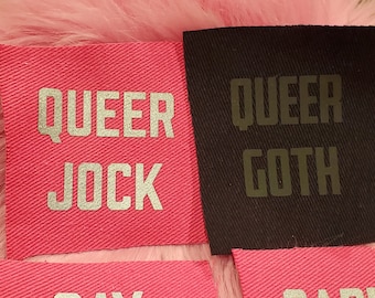 Może przedstawiać: Dwa różowe łatki z tkaniny z srebrnymi napisami "QUEER JOCK" i "QUEER GOTH".