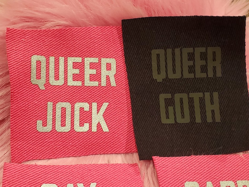 Może przedstawiać: Dwa r&oacute;żowe łatki z tkaniny z srebrnymi napisami "QUEER JOCK" i "QUEER GOTH".