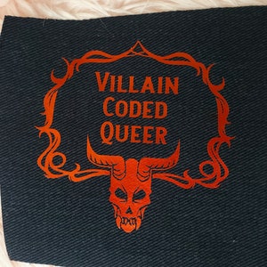 May include: A black fabric patch with an orange design. The design features a skull with horns and a decorative frame with the text "VILLAIN CODED QUEER".