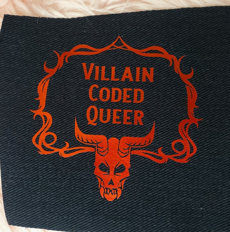 Peut inclure: Un patch en tissu noir avec un motif orange. Le motif repr&eacute;sente un cr&acirc;ne avec des cornes et un cadre d&eacute;coratif avec le texte "VILLAIN CODED QUEER".