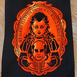 May include: Orange embroidered patch with a black background. The patch features a woman with long dark hair holding a man's head in her hands. The text around the image reads "We gladly feast on those who would subdue us."